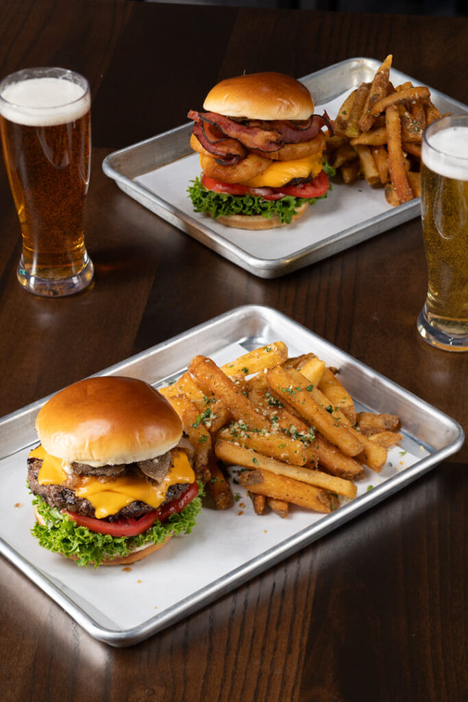 Two burgers served with fries on metal trays with glasses of beer on a wooden table