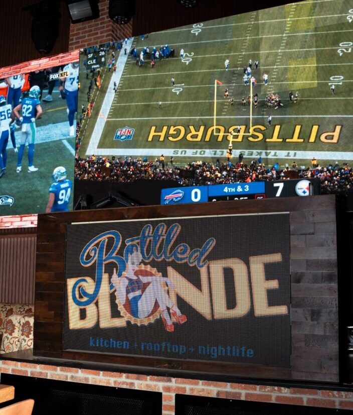 Interior view of a bar featuring a large video wall showing a football game above a branded wall sign