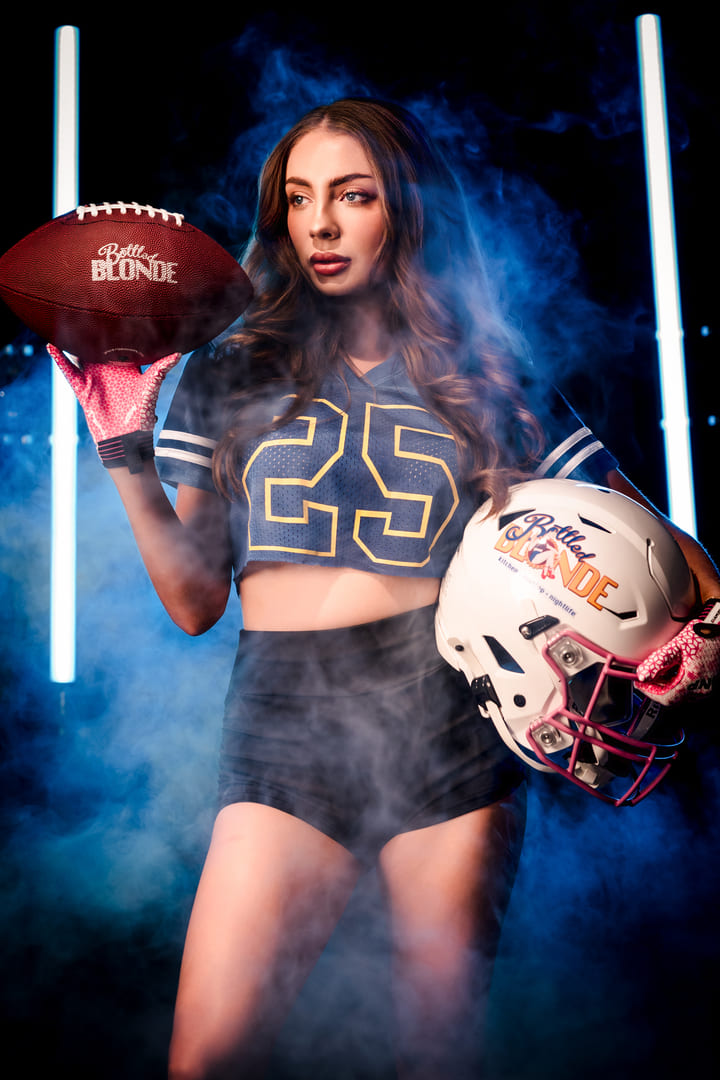 Person wearing a sports jersey holding a football and helmet, standing in a studio setting with dramatic lighting and smoke
