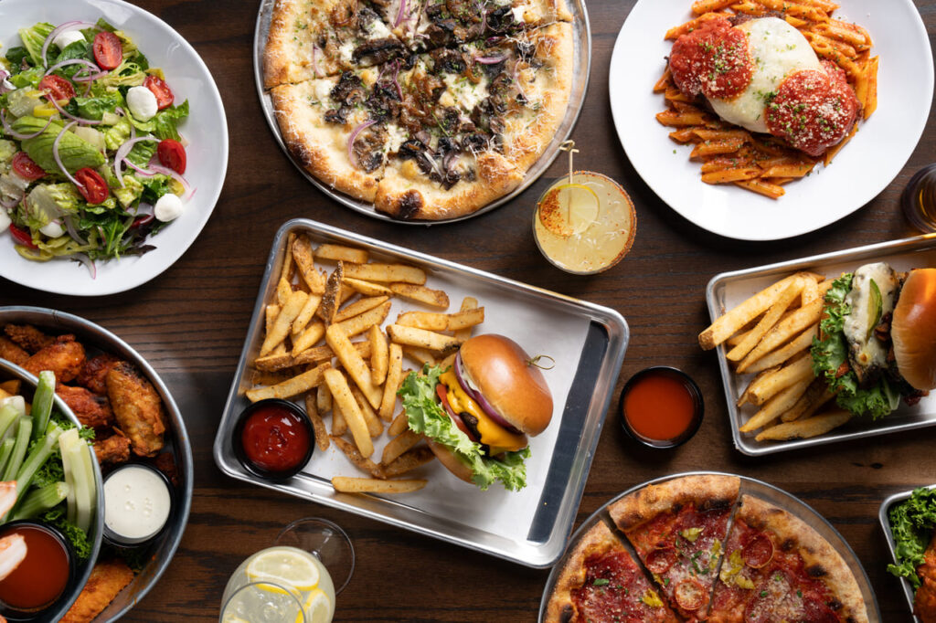 Overhead view of a restaurant table with pizza, burgers with fries, pasta dishes, salad, and chicken wings