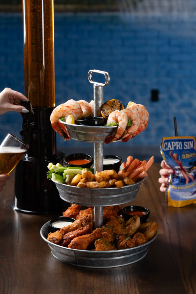 Three-tier appetizer platter with shrimp, chicken wings, fries, vegetables, and dipping sauces on a table