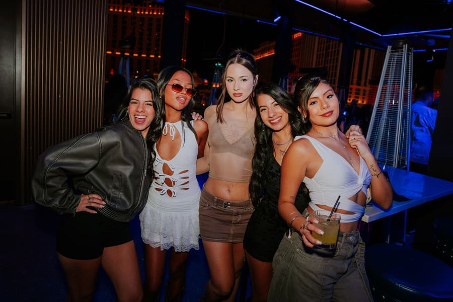 Group of friends posing together at a nightclub patio with drinks under neon lighting.