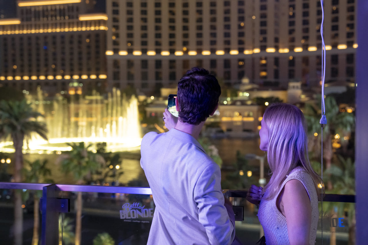 Couple taking a photo of the Bellagio fountains at night from a balcony overlooking the Las Vegas Strip.