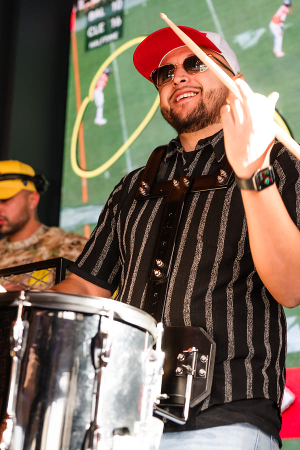 Smiling drummer wearing a red cap and sunglasses playing a drum with a large sports screen in the background.