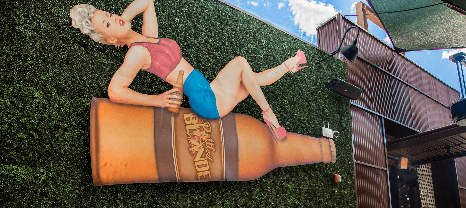 Vintage pin-up style mural of a woman sitting on a giant beer bottle displayed on an outdoor patio wall at Bottled Blonde.